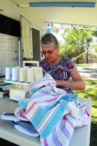 A lady sits at a table outside her caravan. She is sewing striped fabrics on her overlocker.