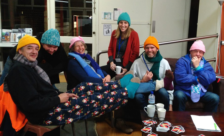 A group of middle aged people dressed in winter pyjamas and beanies sit around playing card games and drinking hot drinks.