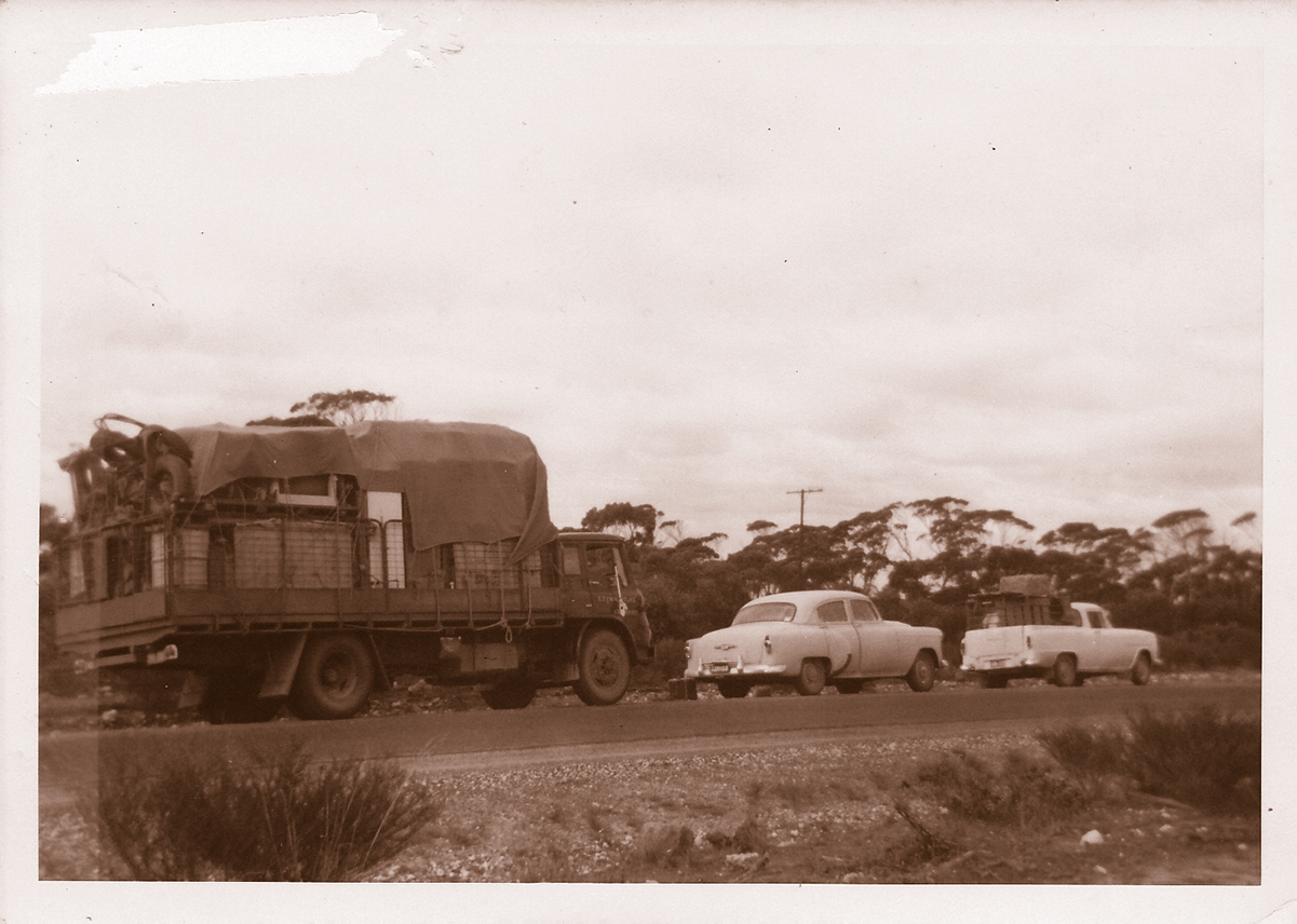 Two vintage cars and a vintage truck are pictured parked on the side of a country road. The truck is laden with furniture and boxes. The photo is sepia toned and is aged. 